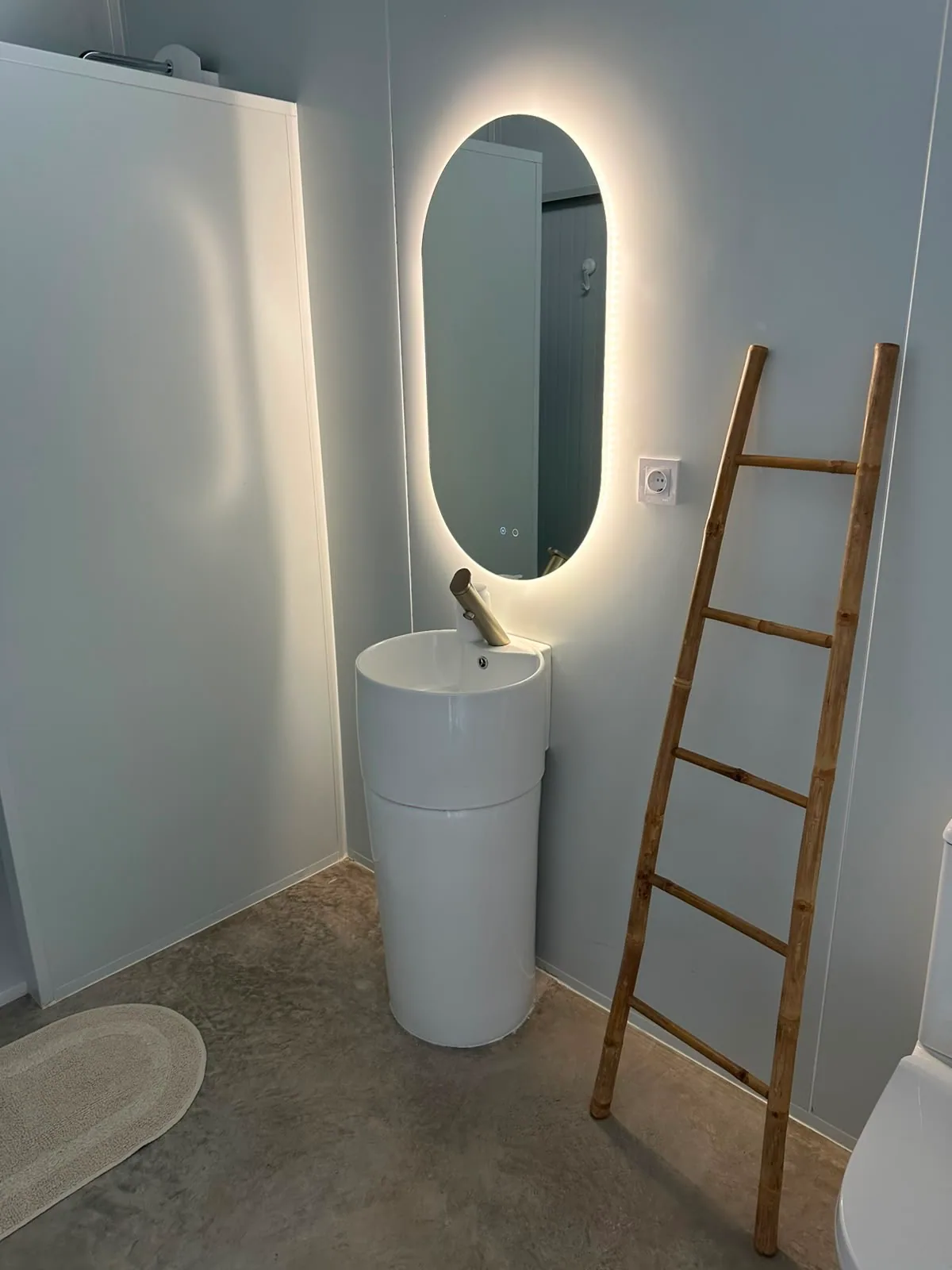 Baño de tonos claros con ducha, lavabo cilíndrico y espejo retroiluminado